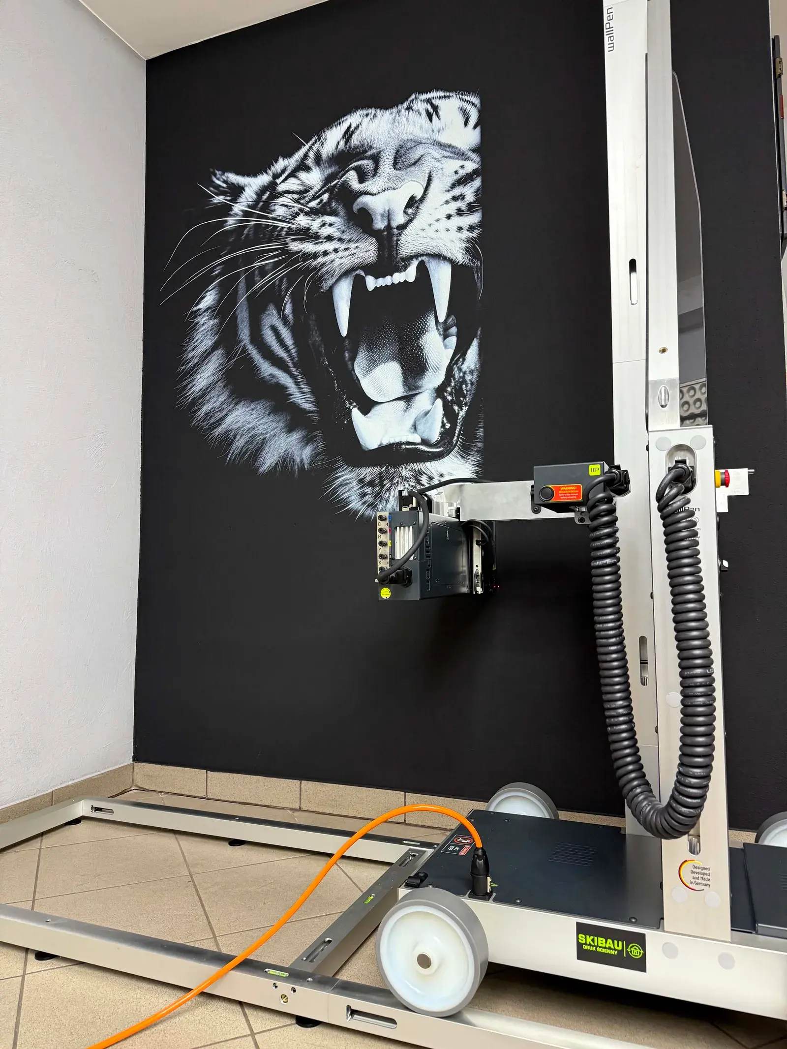 Fotorealistyczny wydruk białego tygrysa na czarnej ścianie wykonany drukarką wallPen, widoczne urządzenie drukujące w trakcie pracy